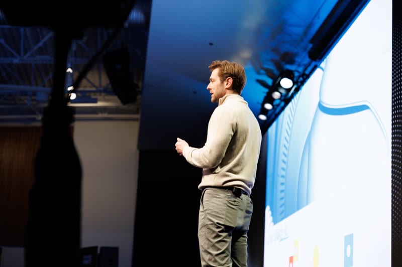 A speaker on stage in front of a bright slide, under blue stage light