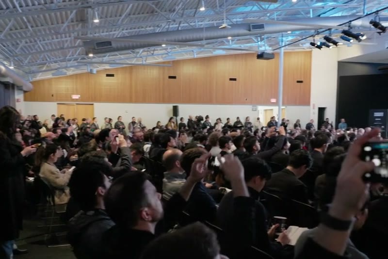 A packed hall with attendees filming the stage on their phones