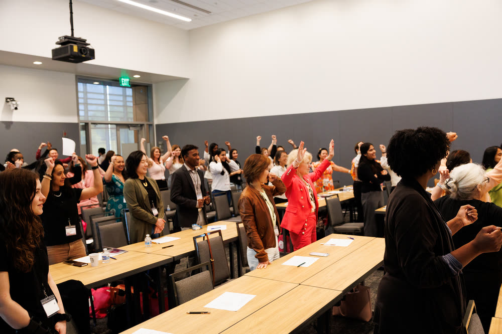 A full room of attendees at round tables raising their hands to answer the speaker