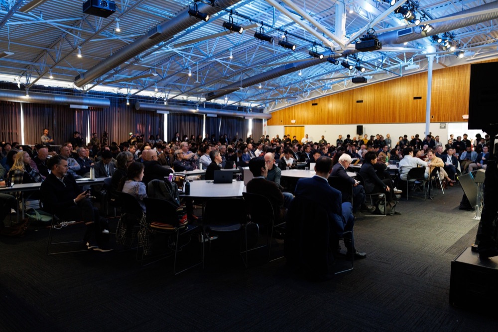 A wide view of a hall with hundreds of seated attendees under truss lighting