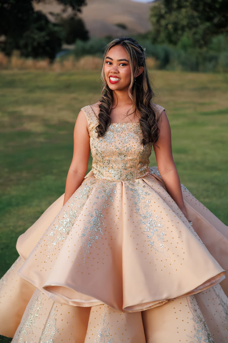 A young woman in a full gold ball gown standing on a lawn, smiling