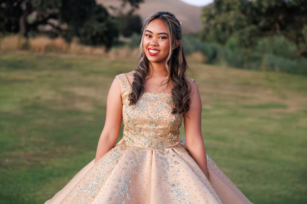 A young woman in a gold ball gown smiling on a lawn at golden hour