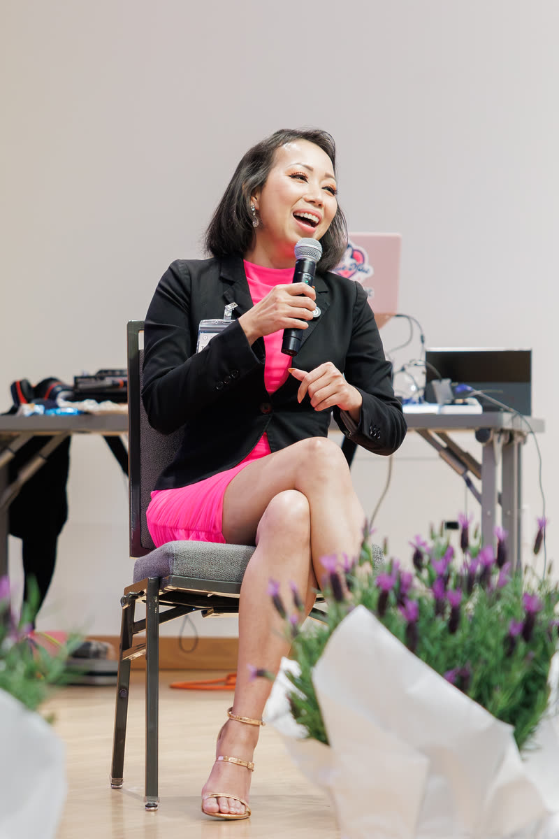 A speaker in a black blazer and pink skirt seated on stage, holding a mic