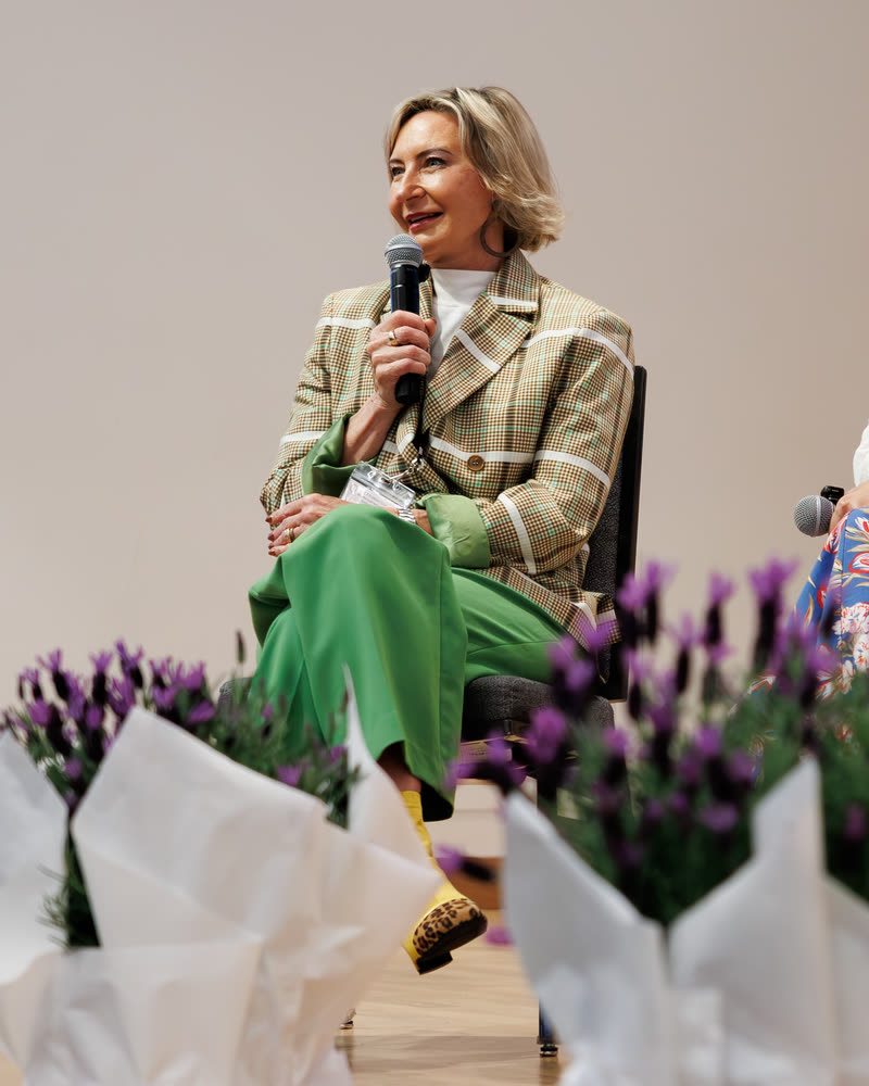 A speaker in a plaid blazer laughing into a mic, framed by lavender arrangements