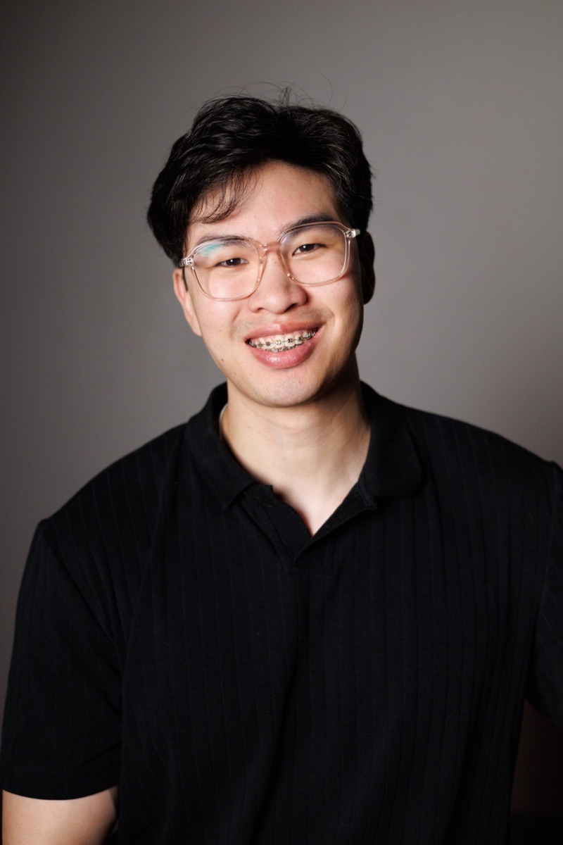 Studio headshot of a young man in glasses, smiling, on a gray background