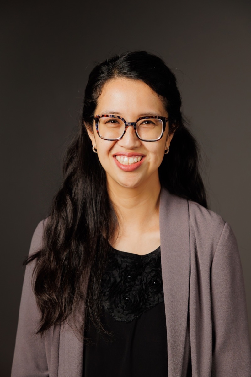 Studio headshot of a woman in glasses, smiling, on a dark background