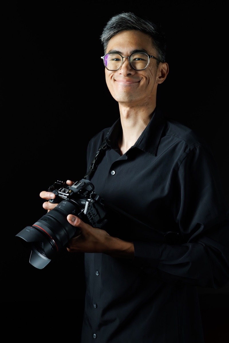 Kyle smiling and holding his camera, photographed on a black background