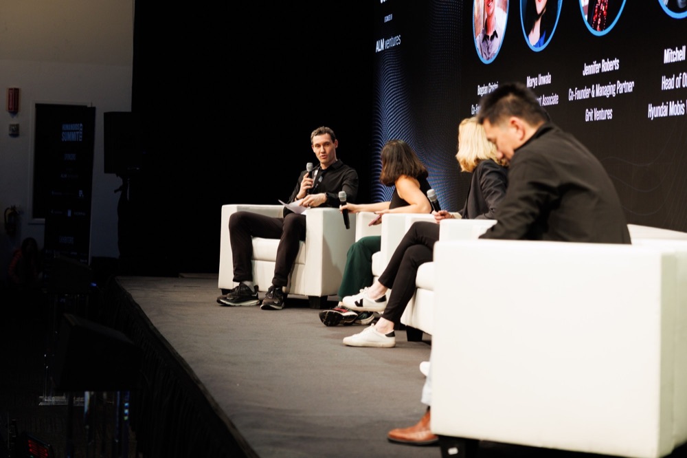 A panel of four on white couches on a dark stage, with speaker headshots on the screen behind them