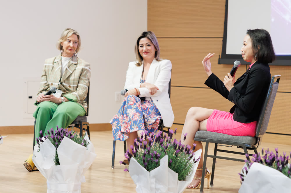 Three women in conversation on stage chairs, framed by lavender arrangements