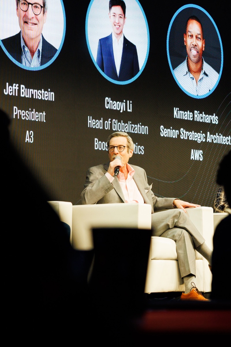 A panelist speaking into a microphone under a screen listing speaker names