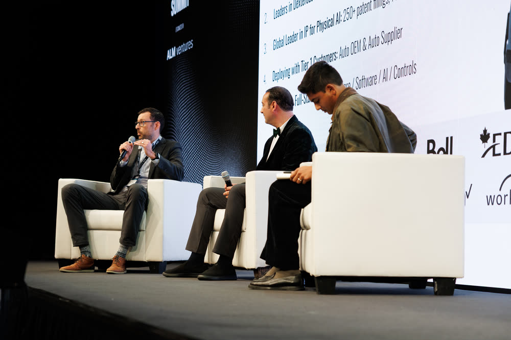 Three panelists on white chairs on a dark stage, one speaking into a mic