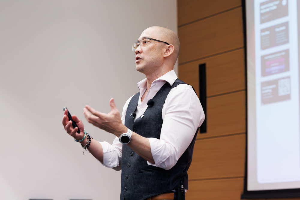 A speaker in a vest gesturing mid-talk, holding a slide clicker