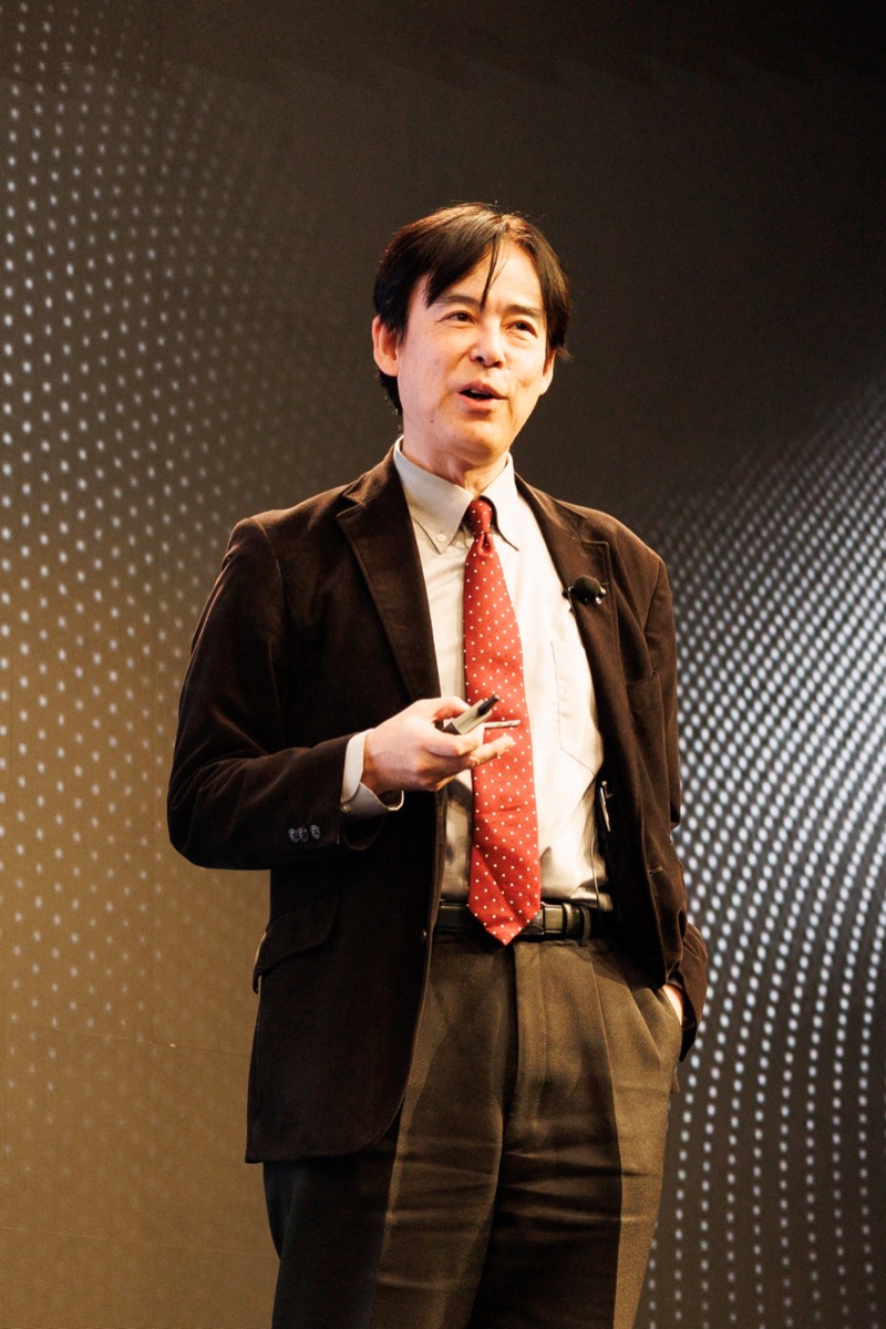 A speaker in a brown jacket and red tie, in warm light against a dotted backdrop
