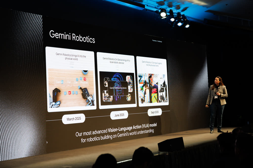 A speaker on a dark stage beside a large Gemini Robotics slide
