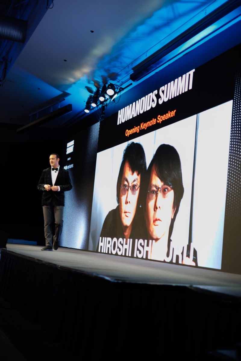 The opening keynote speaker on a dark stage beside a large screen reading Humanoids Summit