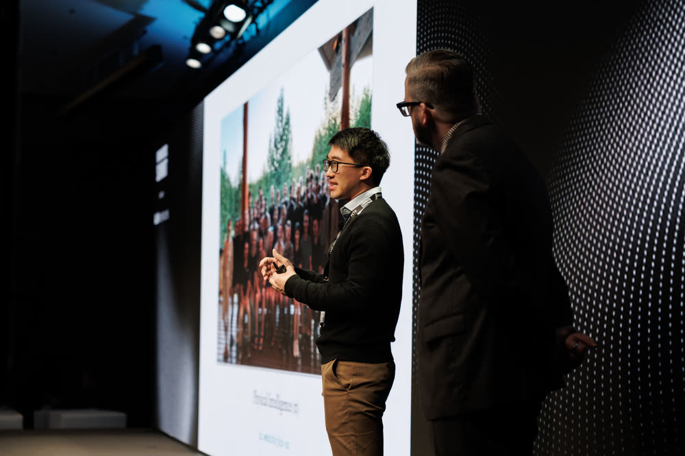 A speaker on stage beside a giant screen showing a crowd photo