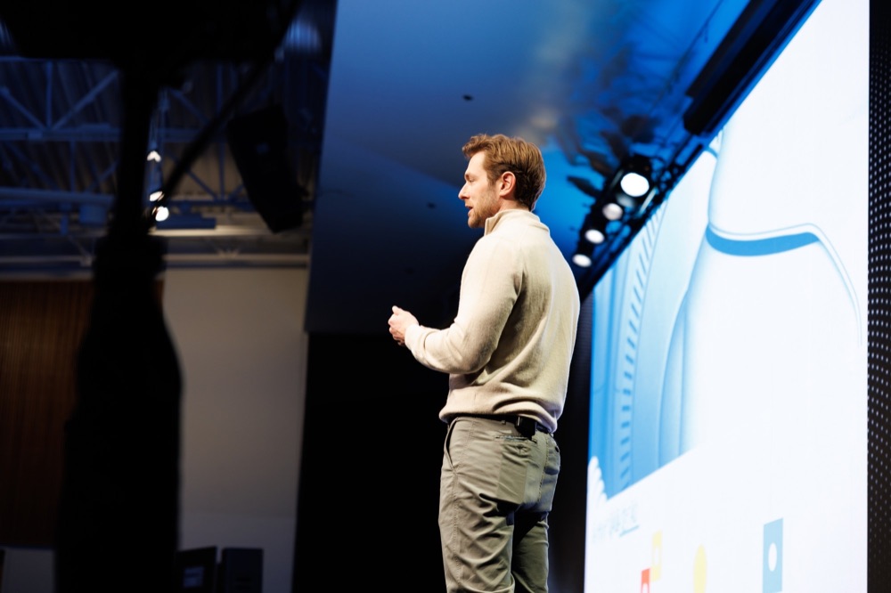 A speaker on stage in front of a bright slide, under blue stage light