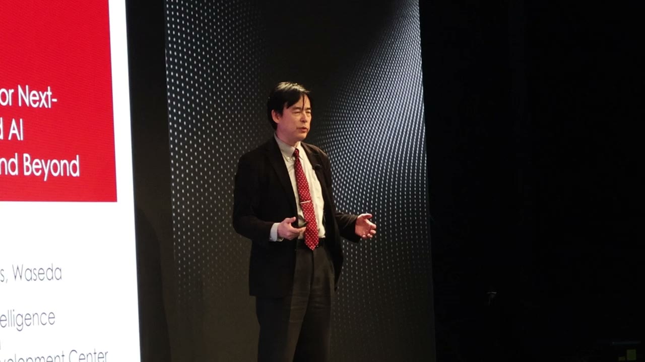 A presenter on a dark stage in front of a slide